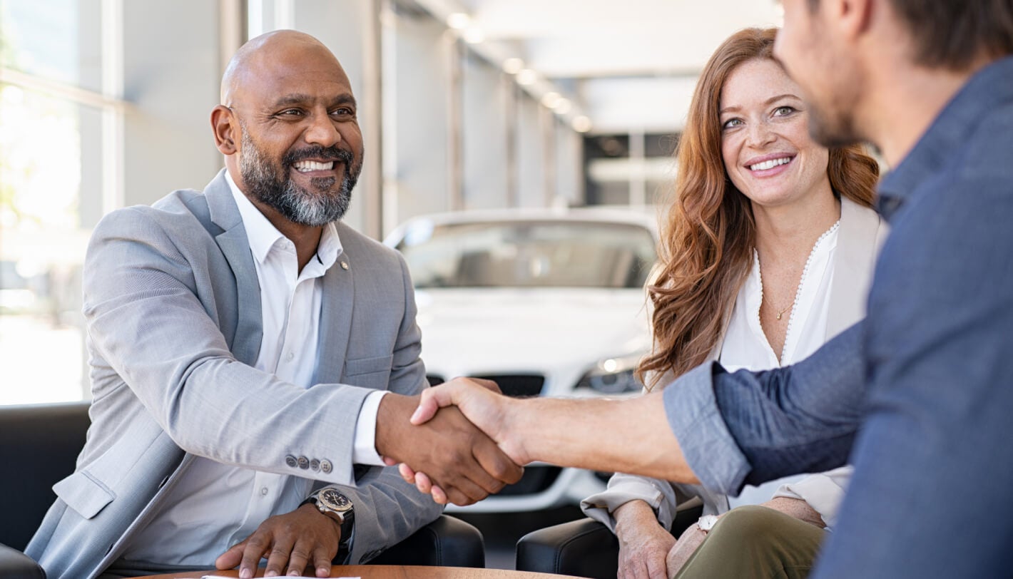 Car salesman shaking hands with smiling customer in a dealership showroom.