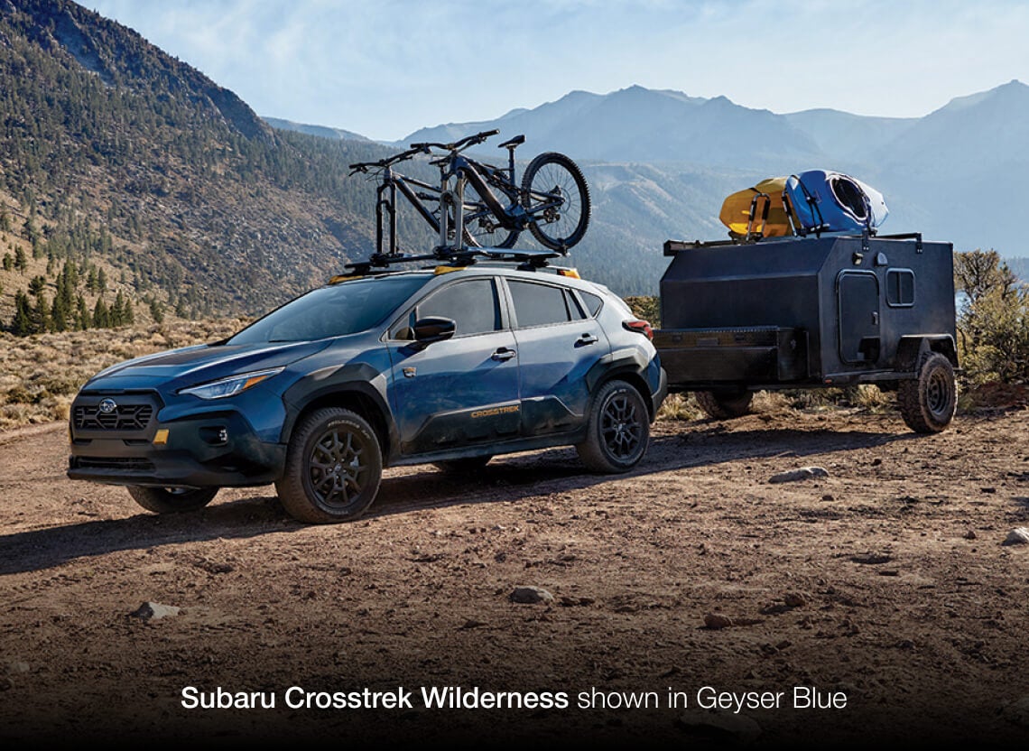 2026 Subaru Crosstrek Wilderness in Geyser Blue, towing an adventure trailer on a rugged mountain trail.