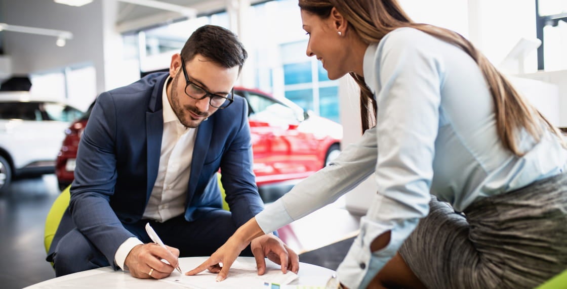 Sales representative and customer reviewing and signing paperwork together inside a car dealership showroom.