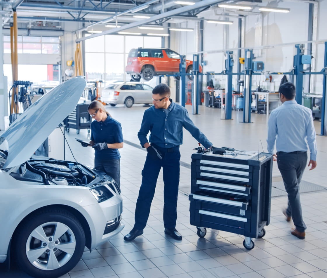 Service technicians inspecting a vehicle with its hood open inside a dealership service center.